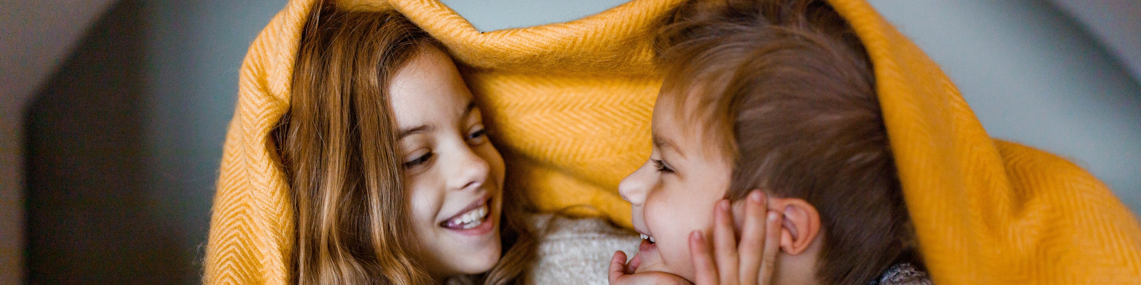 Ein junges Mädchen und ihr kleiner Bruder liegen unter einer gelben Wolldecke und lächeln sich an | © BraunS - Getty Images/iStockphoto
